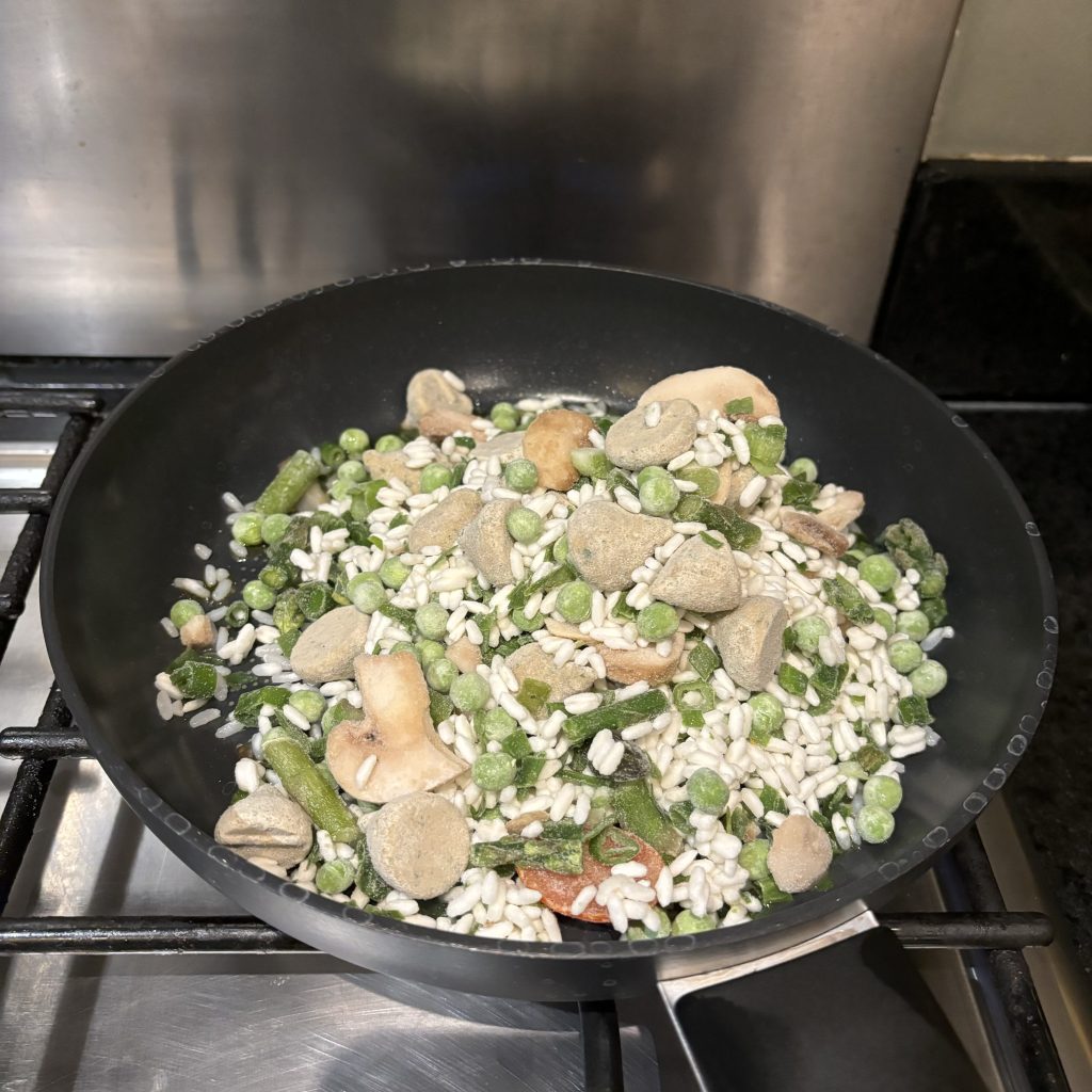 Frying pan on a hob with the frozen contents of a Lazy Vegan Italian risotto meal emptied into it. Rice, peas, beans, mushrooms and sauce frozen into Hersheys Kisses-style lumps.