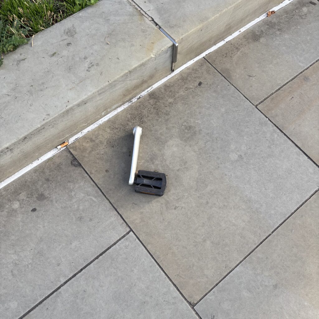 A bicycle pedal and crank, sitting abandoned on the pavement.